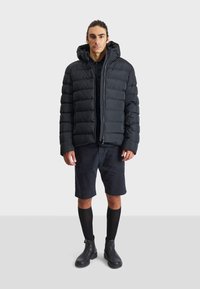 Black puffer jacket with a hood, featuring quilted texture and front zipper. Worn over a black shirt, paired with dark shorts and black boots.