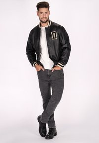 Veste varsity en cuir noir avec des poignets rayés, ornée d'un patch en forme de lettre "D". Portée sur une chemise blanche et associée à un jean sombre et des bottes noires.