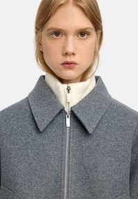 Szara wełniana kurtka z kołnierzem i zamkiem z przodu. Pod nią znajduje się sweter z wysokim kołnierzem w kolorze kremowym. Gładka tekstura.