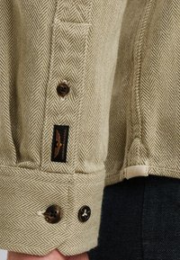 Manche de chemise à motif en chevrons en tissu beige, comportant deux boutons marron et une étiquette noire avec du texte. Design texturé et ajusté.