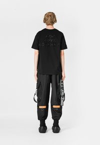 Camiseta negra con el texto en relieve "MOD WAVE MOVEMENT" en la parte de atrás, combinada con pantalones cargo negros que tienen múltiples bolsillos y acentos naranjas.