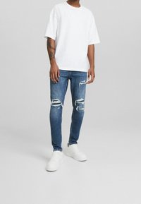 Biała bawełniana koszulka o luźnym kroju, zestawiona z niebieskimi dżinsami denimowymi z postarzanymi detalami i rozdarciami na kolanach, noszona z białymi trampkami.