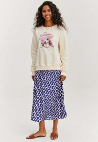 Krémová mikina s ilustrovanou pláží a textem, spárovaná s modrou midi sukni s vlnkovaným vzorem a hnědými šlapkami.