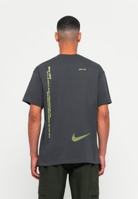 Mand iført en mørkegrå Nike T-shirt med gule bogstaver, der opremser byer, og en prikket Nike swoosh på ryggen, tonet med mørkegrønne cargo-bukser.