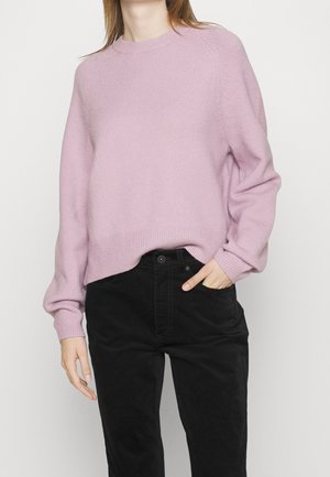 Femme portant un pull à manches longues en maille violet clair et un pantalon taille haute noir, avec une main dans la poche, sur un fond uni.