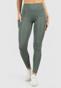 Žena v olivově zelených legínách s vysokým pasem a bočními kapsami, obutá v bílých sportovních botách, stojící na jednotném pozadí.
