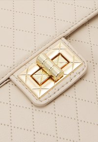 Beżowa pikowana skórzana torba z prostokątnym złotym zapięciem na obracany zatrzask, ozdobionym wygrawerowanymi krzyżami i cylindrycznym obrotowym prętem.