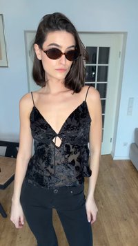 Femme portant un haut sans manches en velours noir à fleurs, un pantalon noir et des lunettes de soleil marron, debout à l'intérieur près d'une porte blanche avec des panneaux en verre.