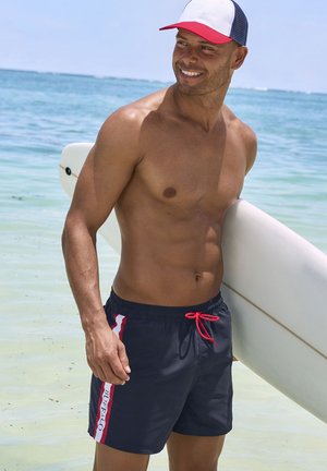 Marineblaue Badehosen mit ropem Kordelzug, die vertikale weiße und rote Streifen an den Seiten aufweisen. Ein weißes Surfbrett liegt neben ihm.