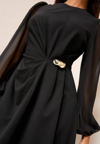 Schwarzes Kleid mit fließenden, transparenten Ärmeln, einem verdrehten Oberteil und einem dekorativen goldfarbenen Knopf, der die Taille betont.