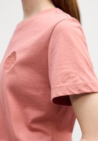 Rosa bomulls t-skjorte med rund hals, korte ermer, med brodert logo på brystet og ermet. Glatt tekstur.