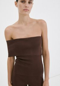 Žena v hnedom prsnom vrchu s odhalenými ramenami a rebrovanom úpletu a v zodpovedajúcich nohaviciach na bielom pozadí.