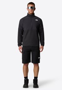 Zwarte fleecetrekker met een kwart-rits en hoge kraag, gecombineerd met zwarte cargoshorts met zijzakken en een logo. Zwarte laarzen en een zonnebril.