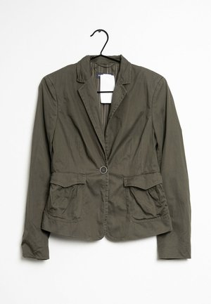 Chaqueta verde oliva con cuello de muesca, cierre de un solo botón, dos bolsillos frontales y tejido texturizado, colgando de un gancho negro.