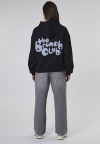 Felpa nera oversize con la scritta "the brunch club" in grigio chiaro. Abbinata a jeans grigi a gamba larga e sneakers color crema.