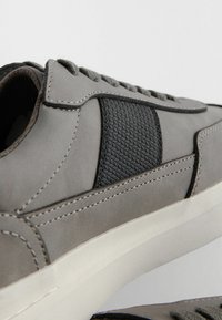 Sneaker grigio con una tomaia in pelle liscia, pannello elastico nero testurizzato, cuciture a contrasto e suola in gomma bianca.