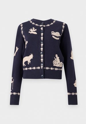 Un mannequin se tient devant un fond de studio blanc, vêtu d’un cardigan bleu marine à motifs crème, d’un jean bleu à jambes larges et de chaussures noires.