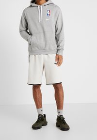 Sweat à capuche gris avec le logo NBA et des cordons blancs, associé à un short crème avec un bord noir, et des chaussures de sport vert foncé.