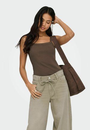 Mujer con cabello largo y oscuro, vistiendo una camiseta sin mangas marrón y pantalones anchos beige con cordón, llevando un gran bolso marrón al hombro, posando con la mano en la cabeza.