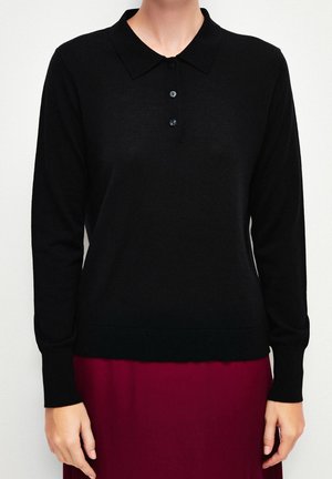 Chemise noire à manches longues avec col et trois boutons sur le devant, portée sur une jupe rouge, montrant le torse et les bras.