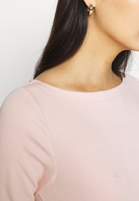 Helles rosa Sweatshirt mit einem Bootsausschnitt und glatter Textur, kombiniert mit runden Ohrringen aus Goldtone mit dunklem Akzent.