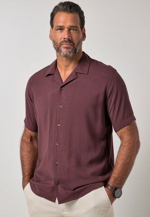 Man met kort donker haar en baard, draagt een bordeauxkleurig kortermig overhemd met knopen en lichte broek, handen in de zakken.
