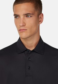Črna polo majica s klasičnim ovratnikom, tri gumbe na zapenjanju in gladko, lahko teksturo. Preprosta zasnova brez vzorcev ali logotipov.