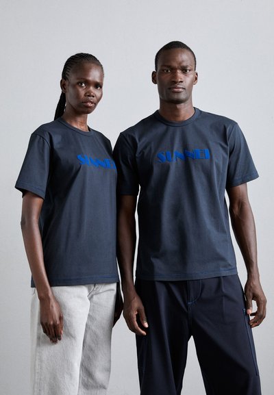 Navy t-shirts with blue "SUNNEI" logo at the center, short sleeves, and round neck. Worn with gray jeans and dark trousers. Simple design.