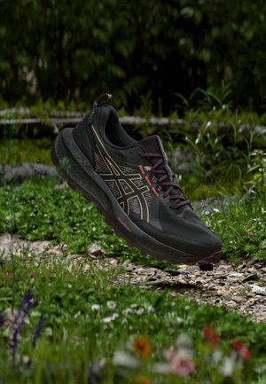 Chaussure de course noire et violette avec une semelle à motifs flottant au-dessus d'un chemin de gravier entouré de plantes vertes et de fleurs sauvages.