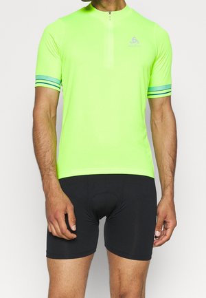 Mann trägt neongelbes kurzärmeliges Fahrradtrikot mit Frontreißverschluss und schwarze, gepolsterte Fahrradshorts.