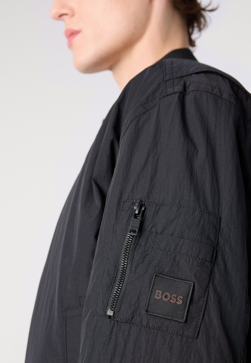 Czarna, lekka kurtka charakteryzuje się teksturowanym materiałem, boczną kieszenią na zamek błyskawiczny oraz kontrastową naszywką z logo. Dopasowany krój z wyraźnym kołnierzem.