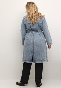 Blondýnka stojí k nám zády, oblečená v doplněném denimovém trenčkotě, černých kalhotách a černých plochých botách, na pozadí světle jednobarevné.