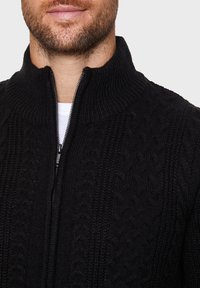 Pull noir à fermeture éclair, présentant un motif torsadé, un col haut et un tissu texturé. La fermeture éclair descend sur le devant pour un ajustement réglable.