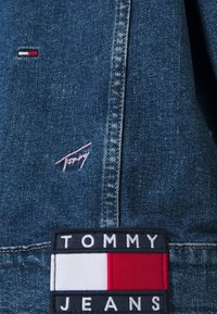 Donkere blauwe denimstof met een roze "Tommy"-logo en een rechthoekig zwart label met "TOMMY JEANS" en een driekleurige accent in rood, wit en blauw.