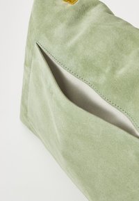 Sac en suède vert présentant une texture lisse et une poche avant visible avec un détail de couture contrastante. Quincaillerie de fermeture en haut.