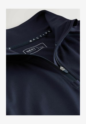 Navy activewear top with half-zip design, soft fabric, flat seams, and a label reading "NEXT ACTIVE." Features a smooth texture and a collar.