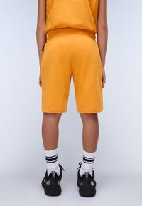 Gula bomullsshorts med elastisk midja, med längd till mitten av låret, med en sidficka och i kombination med svarta sneakers och randiga strumpor.