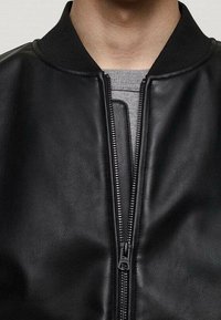 Gros plan sur une personne portant une veste en cuir noire avec une fermeture éclair, par-dessus une chemise grise à col côtelé.