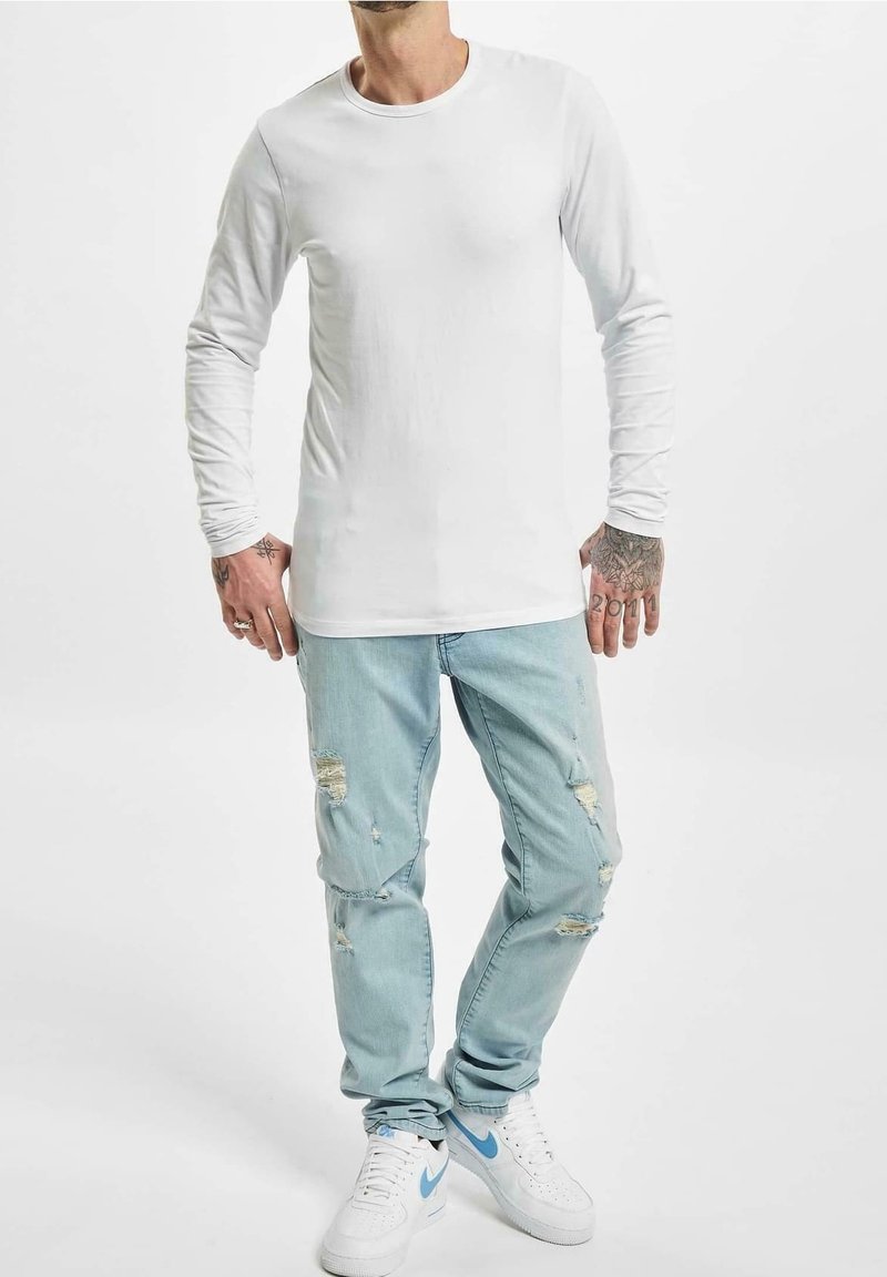 T-shirt bianca a maniche lunghe con collo rotondo, abbinata a jeans chiari strappati e sneakers bianche con dettagli blu.