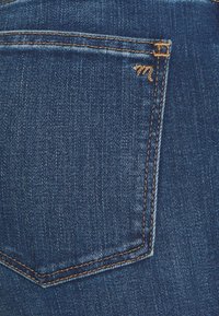 Tecido de denim em azul escuro com um bolso traseiro com costura dourada e um pequeno logótipo bordado na borda. Textura suave e design clássico.