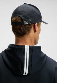 Cappello da baseball blu navy con cinturino regolabile, presenta un accento argento. Felpa con cappuccio nera ha strisce bianche sulla schiena.