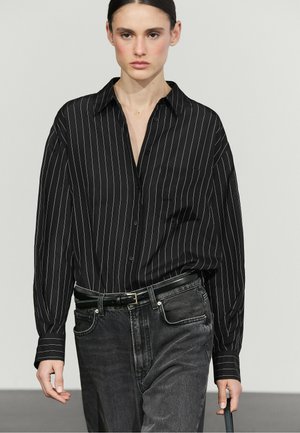 Jeune personne portant une chemise noire à fines rayures boutonnée, rentrée dans un jean gris foncé avec une ceinture noire, debout devant un fond uni.