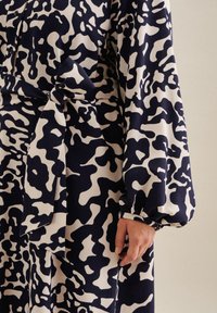 Robe à motifs abstraits bleu marine et beige avec de longues manches ballon, des poignets boutonnés et une ceinture nouée à la taille d'une personne.