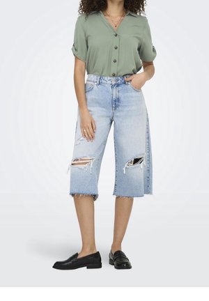 Mujer con shorts vaqueros desgastados azul claro hasta la rodilla, camisa verde salvia de manga corta con botones y mocasines negros sin cordones.