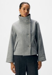 Graue kurze Jacke mit hohem Kragen, ausgestattet mit sechs großen Kn�öpfen, seitlichen Taschen und einem strukturierten Stoff. Die Silhouette ist oversized mit weit geschnittenen Ärmeln.