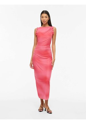 Nő áll, rózsaszín ujjatlan midi ruhát visel, amelynek vállai redőzöttek, és fekete pántos magassarkú szandált visel, fehér háttér előtt.