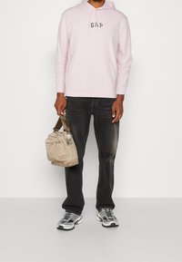 Felpa rosa chiaro con logo "GAP" blu navy, jeans neri, sneakers grigie e borsa beige con finitura testurizzata e tracolla regolabile.