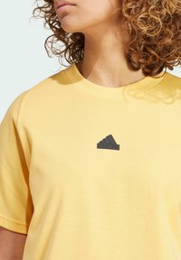 Camiseta de algodón amarilla con cuello redondo y mangas cortas. Presenta un logo de Adidas en caucho negro en el pecho. Textura suave.