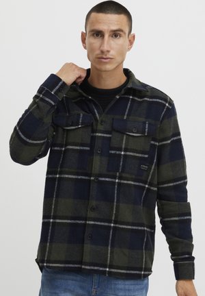 Plaid flannel shirt in navy and olive green, featuring two chest pockets, button closure, and a textured finish.