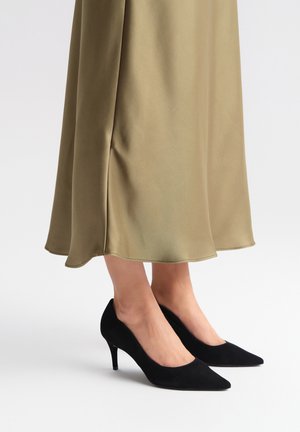 Czarne zamszowe szpilki z wydłużonym noskiem i smukłym obcasem w kształcie szpilki, uzupełnione satynową spódnicą do kolan w oliwkowym odcieniu.